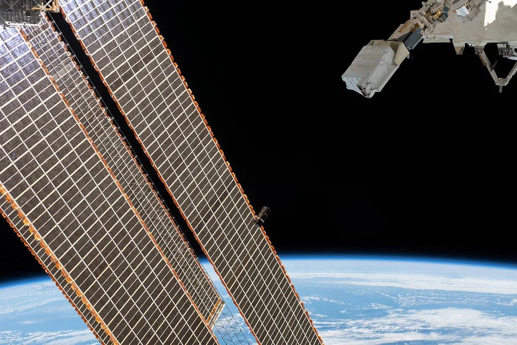 A small CubeSat passing the International Space Station's solar arrays above Earth